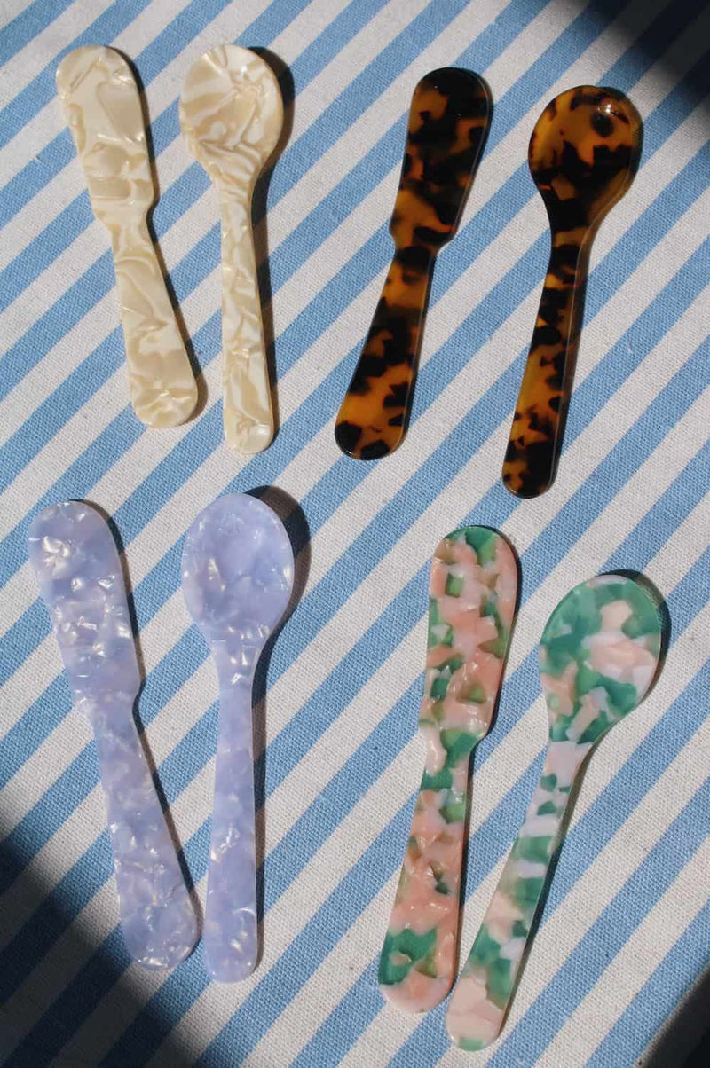 Six decorative plastic spoons with marbled patterns in cream, tortoiseshell, lavender, and green-pink, arranged on a blue and white striped fabric background.