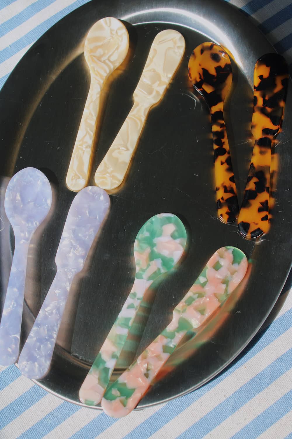 Six decorative plastic spoons in various marbled and patterned designs, including tortoiseshell, cream, lavender, green, pink, and blue, arranged on a black oval tray over a blue and white striped surface.