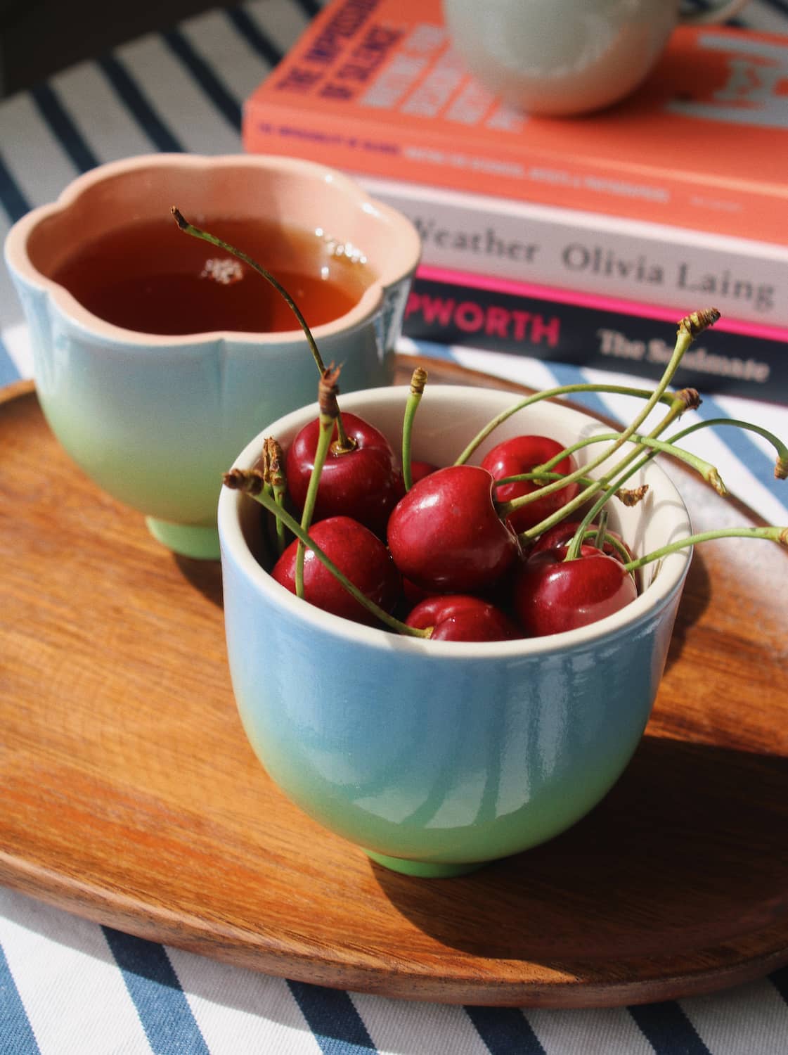 Two ceramic cups with a blue-to-green gradient finish, one filled with tea and the other with red cherries, arranged on a wooden tray with books in the background.