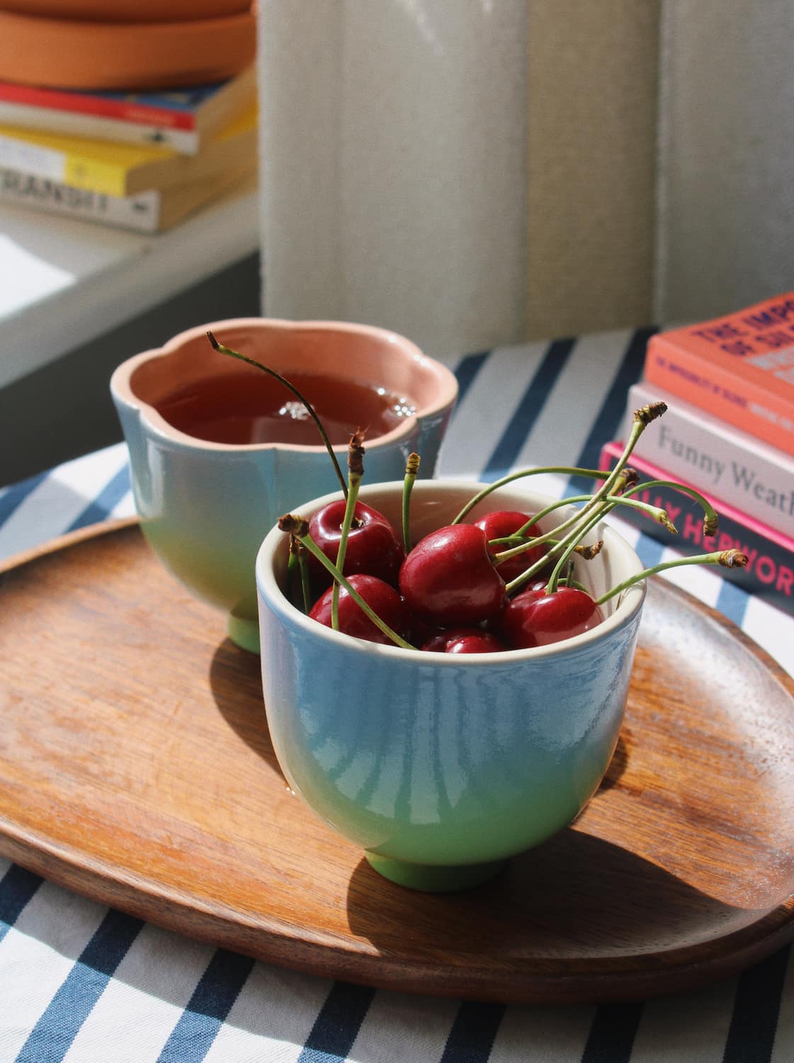 Two ceramic cups with a blue-to-green gradient glaze sit on a wooden tray. One cup is empty, and the other is filled with red cherries.