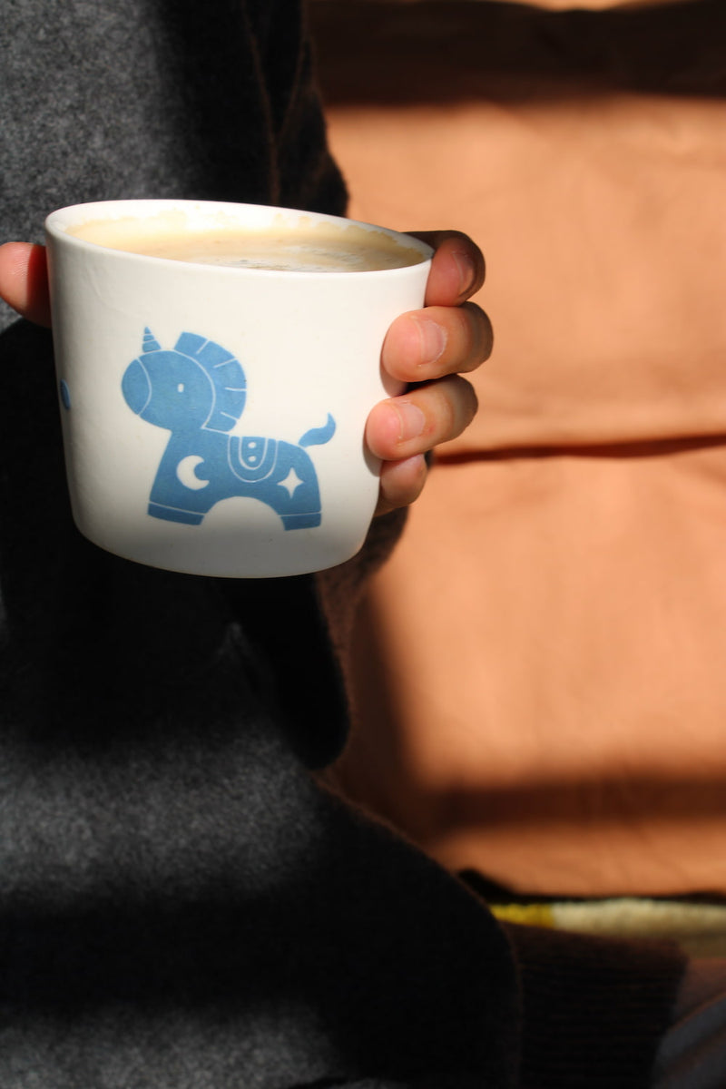 A person holding a white ceramic mug with a blue unicorn graphic, set against a warm brown background with strong sunlight and shadows.