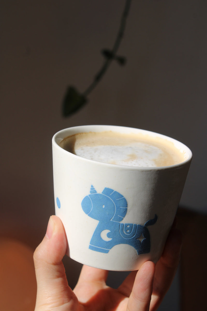 A hand holding a white ceramic cup filled with a latte, decorated with a blue unicorn illustration. The background is a dark, softly lit wall with a plant shadow.