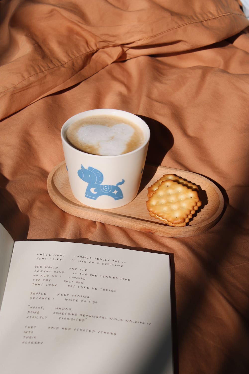A white ceramic cup with a blue cat illustration sits on a wooden tray beside a biscuit, set on rumpled tan fabric with a book in the foreground.