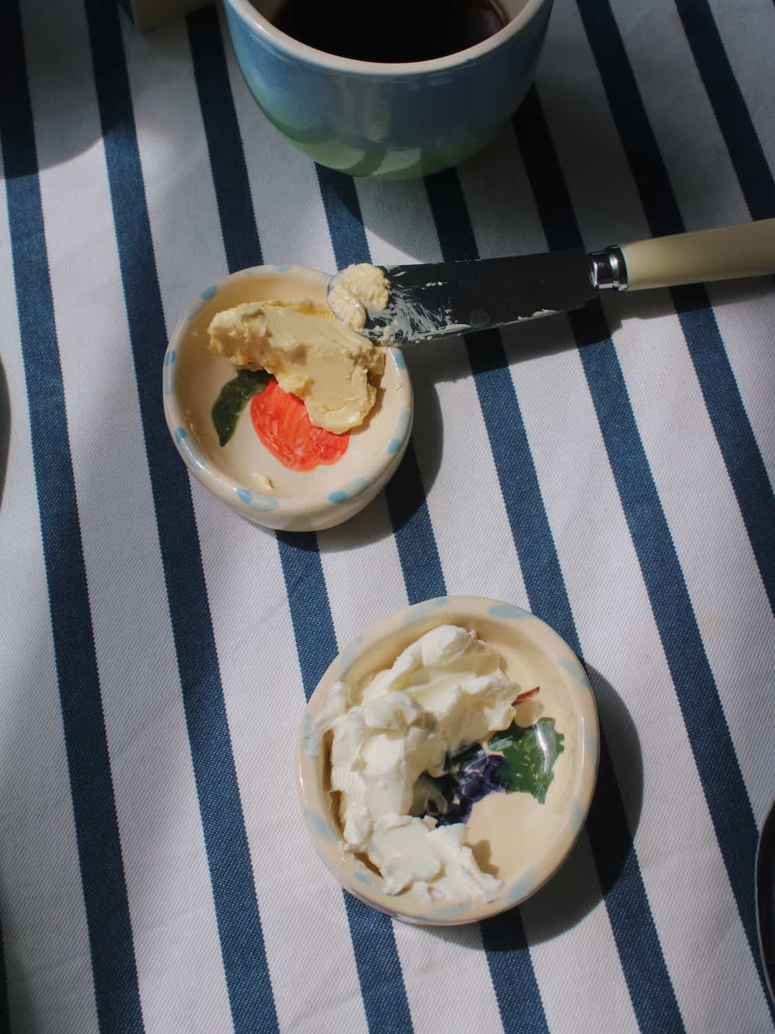 Two small decorative ceramic bowls containing whipped cream and butter, with a knife resting between them on a blue and white striped tablecloth. A ceramic mug is partially visible at the top.