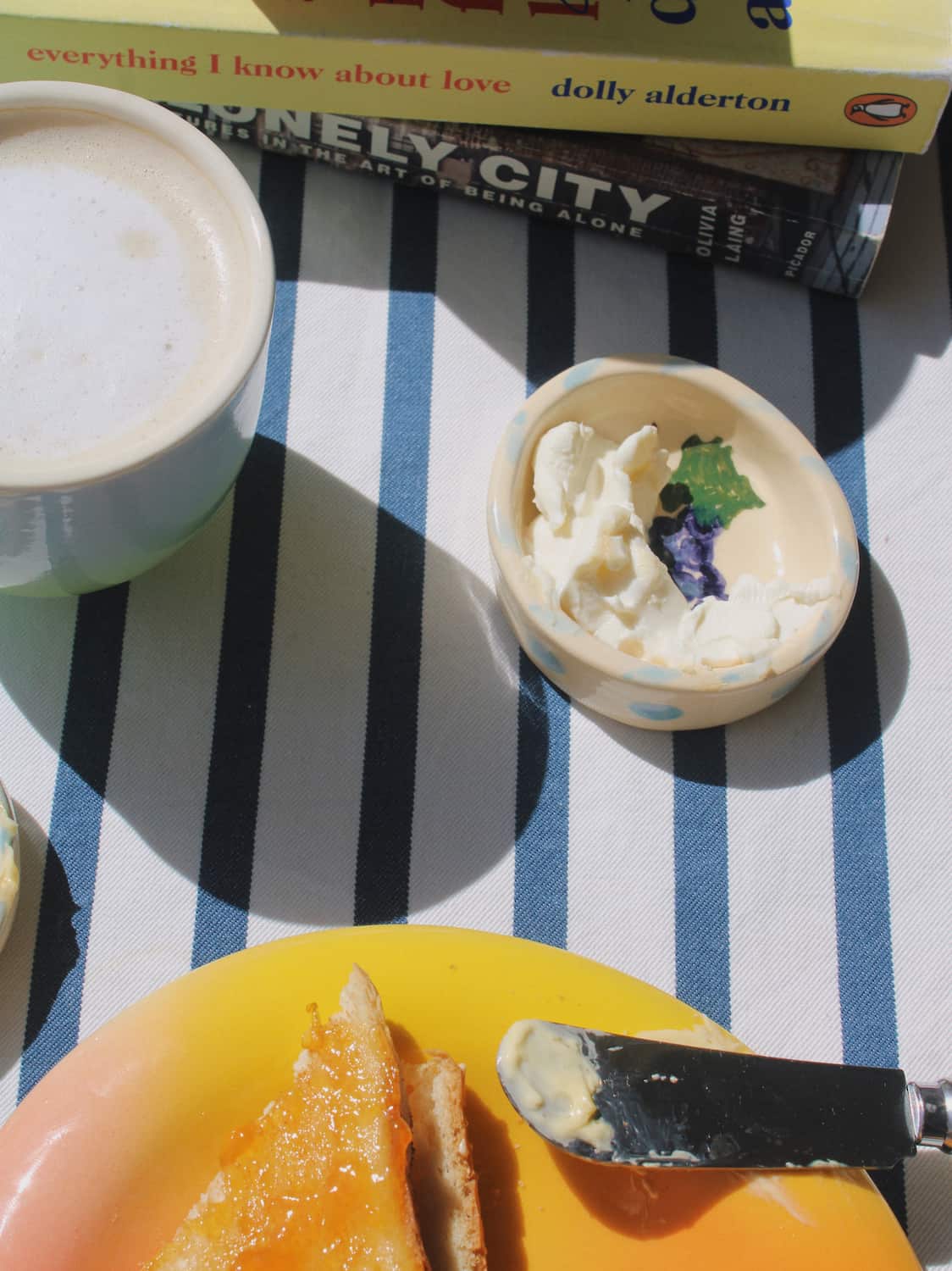 A cozy breakfast scene on a blue and white striped tablecloth, featuring a cup of coffee, a small bowl of clotted cream with a butterfly flower garnish, and a yellow plate with toast and butter in the foreground. Books are stacked in the background.