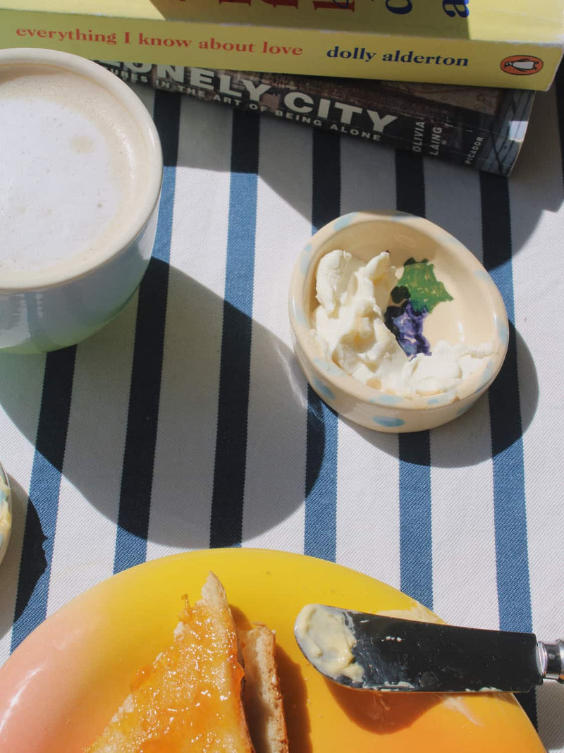 A cozy breakfast scene on a blue and white striped tablecloth, featuring a cup of coffee, a small bowl of clotted cream with a butterfly flower garnish, and a yellow plate with toast and butter in the foreground. Books are stacked in the background.