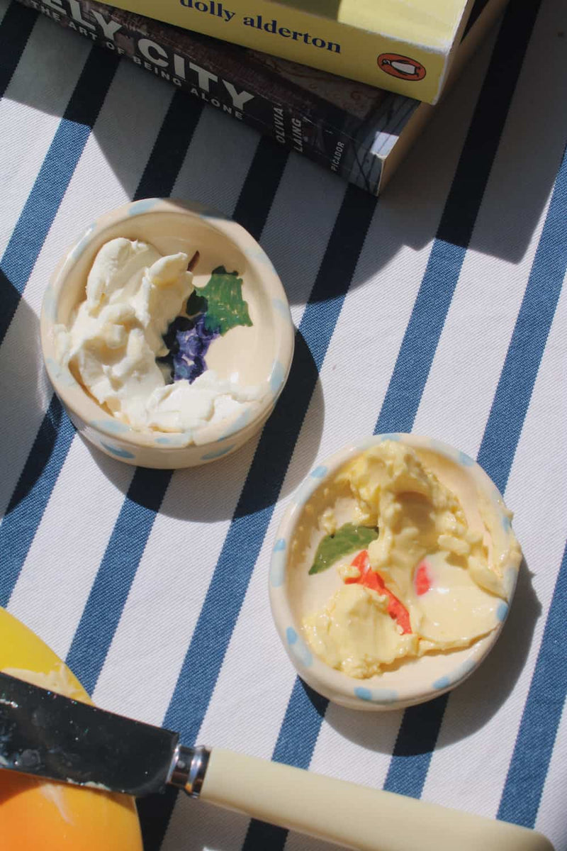 Two small ceramic bowls filled with whipped cream or butter, placed on a blue-and-white striped cloth with a knife and books nearby.